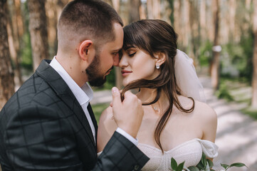 Wedding portrait of newlyweds close up. A bearded, stylish groom in a suit and a beautiful brunette bride in a white dress with a bouquet in her hands are standing and hugging in nature in the forest.