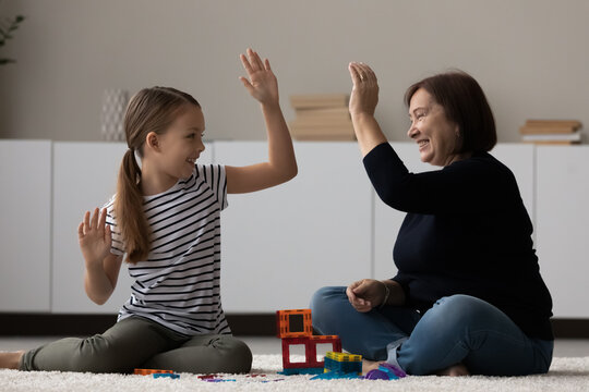 Happy girl and grandma playing on heating floor, constructing toy building, giving high five. Grandmother and granddaughter relaxing, spending time together, enjoying playtime, having fun at home