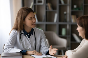 Senior middle aged patient consulting young female doctor at home. Optimistic physician visiting and talking to sick retiree, giving medical help, treatment recommendation, healthcare advice