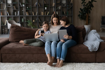 Three female generations of family relaxing on couch with gadgets. Happy kid, mom, grandma using...