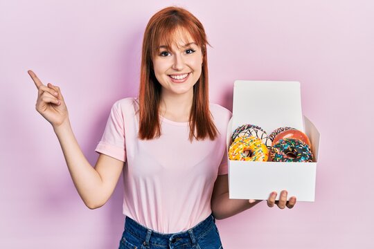 Redhead young woman holding tasty colorful doughnuts smiling happy pointing with hand and finger to the side