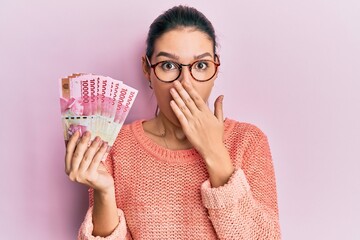 Young caucasian woman holding 100000 indonesian rupiah covering mouth with hand, shocked and afraid for mistake. surprised expression