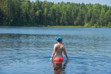 Young woman in a red swimsuit and a blue bandana enters the lake water on a bright summer day