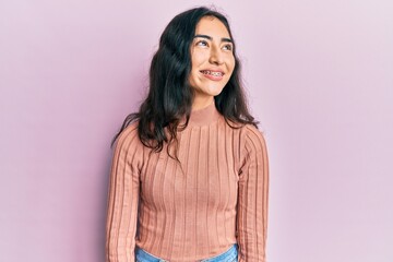 Hispanic teenager girl with dental braces wearing casual clothes looking away to side with smile on face, natural expression. laughing confident.