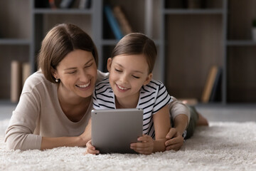 Happy girl and mom relaxing on warm floor, cozy carpet, using tablet, looking at screen, laughing, watching movie, making video call, reading book on internet together. Mother and daughter at home