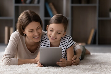 Happy girl and mom relaxing on warm floor, cozy carpet, using tablet, looking at screen, laughing,...