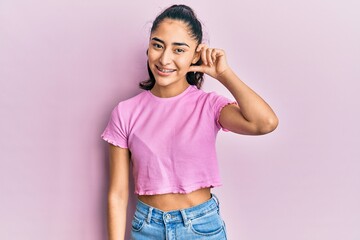 Hispanic teenager girl with dental braces wearing casual clothes smiling with hand over ear listening and hearing to rumor or gossip. deafness concept.