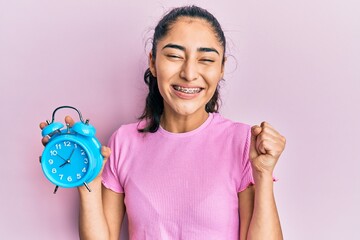 Hispanic teenager with dental braces holding alarm clock screaming proud, celebrating victory and...