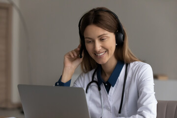 Happy friendly female doctor in headset using laptop, giving online help, medical support, advice,...