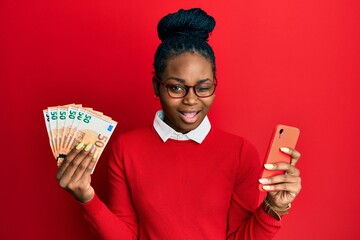 Young african american woman using smartphone holding euro banknotes winking looking at the camera...