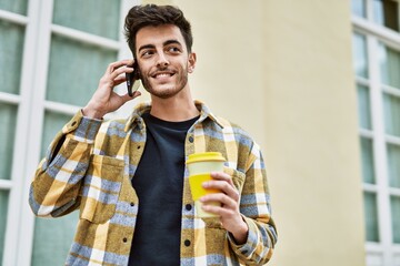 Handsome hispanic man smiling happy and confident at the city speaking on the phone and drinking a cup of coffee