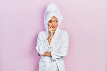 Young hispanic girl wearing shower towel cap and bathrobe serious face thinking about question with hand on chin, thoughtful about confusing idea