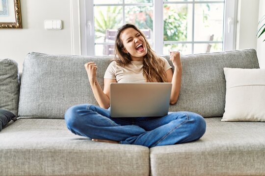 Beautiful Young Brunette Woman Sitting On The Sofa Using Computer Laptop At Home Very Happy And Excited Doing Winner Gesture With Arms Raised, Smiling And Screaming For Success. Celebration Concept.