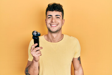 Young hispanic man holding electric razor looking positive and happy standing and smiling with a...