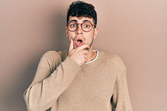 Young hispanic man wearing casual clothes and glasses looking fascinated with disbelief, surprise and amazed expression with hands on chin