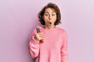 Young brunette woman holding colored pencils scared and amazed with open mouth for surprise,...