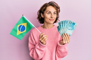 Young brunette woman holding brazil flag and real banknotes smiling looking to the side and staring...