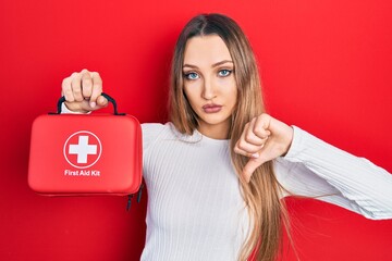 Young blonde girl holding first aid kit with angry face, negative sign showing dislike with thumbs...