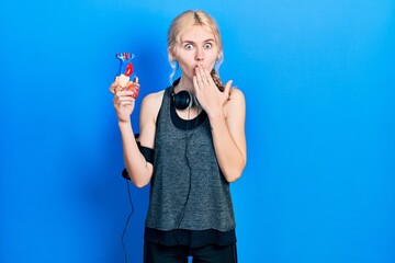 Beautiful caucasian sports woman with blond hair holding heart covering mouth with hand, shocked...