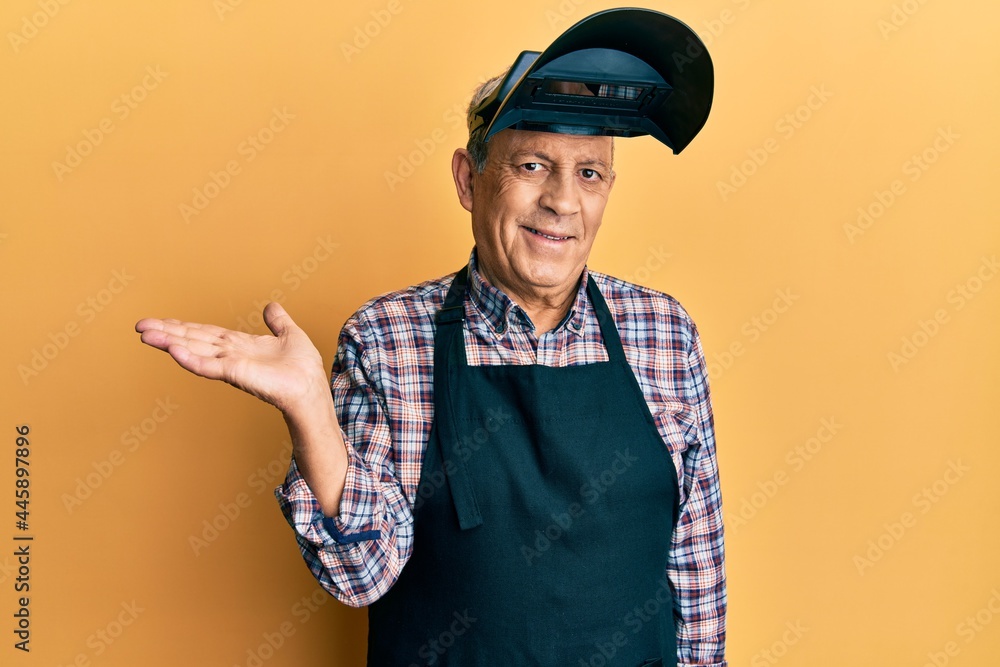 Sticker Handsome senior man with grey hair wearing welding protection mask smiling cheerful presenting and pointing with palm of hand looking at the camera.