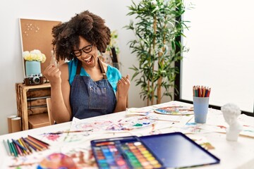 Beautiful african american woman with afro hair painting at art studio very happy and excited doing winner gesture with arms raised, smiling and screaming for success. celebration concept.