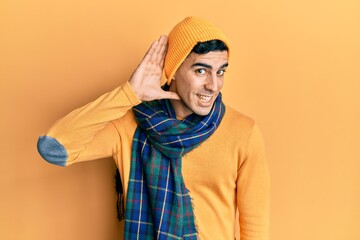 Handsome hispanic man wearing wool hat and winter scarf smiling with hand over ear listening an hearing to rumor or gossip. deafness concept.