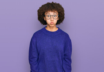 Young hispanic girl wearing casual winter sweater and glasses puffing cheeks with funny face. mouth inflated with air, crazy expression.