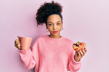 Young african american girl drinking a cup of coffee and eating sweet relaxed with serious...