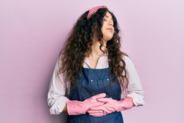 Young brunette woman with curly hair wearing cleaner apron and gloves with hand on stomach because nausea, painful disease feeling unwell. ache concept.