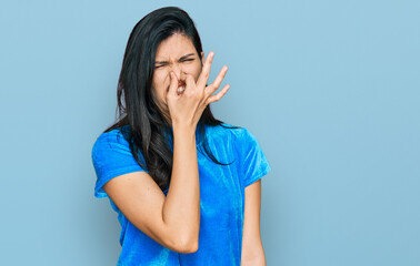 Young hispanic woman wearing casual clothes smelling something stinky and disgusting, intolerable smell, holding breath with fingers on nose. bad smell