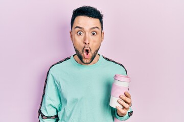 Young hispanic man drinking a cup of coffee scared and amazed with open mouth for surprise,...