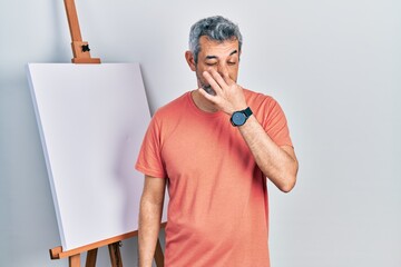 Handsome middle age man with grey hair standing by painter easel stand smelling something stinky...