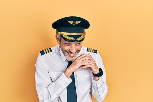 Handsome middle age man with grey hair wearing airplane pilot uniform laughing nervous and excited with hands on chin looking to the side