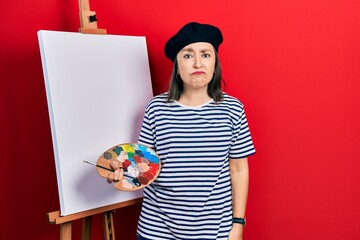 Middle age hispanic woman standing drawing with palette by painter easel stand depressed and worry...