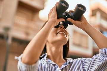Fototapeta premium Young hispanic girl smiling happy using binoculars at the city.