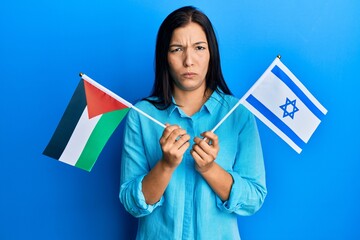 Young latin woman holding palestine and israel flags skeptic and nervous, frowning upset because of...