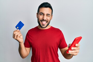 Young hispanic man holding smartphone and credit card sticking tongue out happy with funny expression.