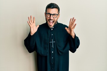 Young hispanic man wearing priest uniform standing over white background celebrating mad and crazy...