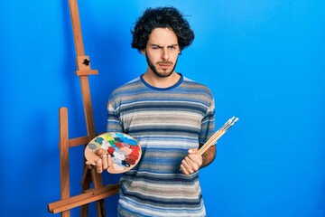 Handsome hispanic man standing drawing with palette by painter easel stand skeptic and nervous,...