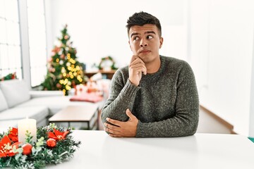 Young hispanic man sitting on the table by christmas tree with hand on chin thinking about question, pensive expression. smiling with thoughtful face. doubt concept.
