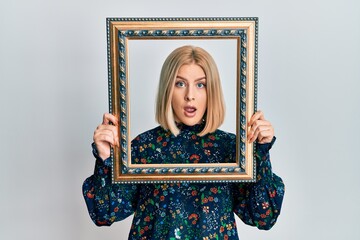 Young blonde woman holding empty frame in shock face, looking skeptical and sarcastic, surprised with open mouth
