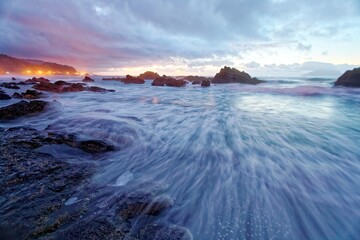 Sunrise scenery of beautiful Wai'ao Beach, in Yilan (Ilan), Taiwan, with turbulent waves rushing upon the rocks & an island on the horizon under moody cloudy sky reflected on sea water (Long Exposure)