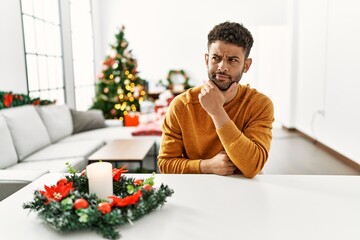 Arab young man sitting on the table by christmas tree with hand on chin thinking about question, pensive expression. smiling with thoughtful face. doubt concept.