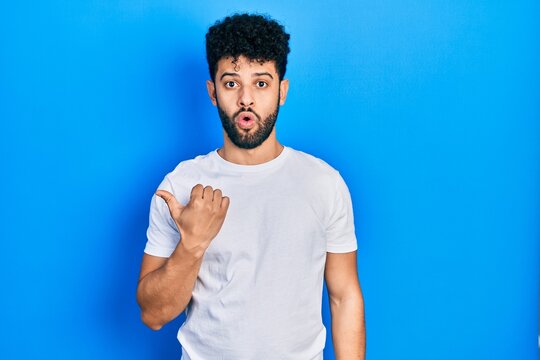 Young arab man with beard wearing casual white t shirt surprised pointing with hand finger to the side, open mouth amazed expression.