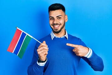 Young hispanic man with beard holding gambia flag smiling happy pointing with hand and finger