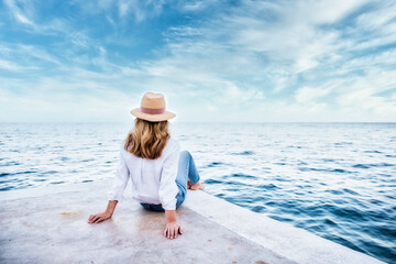 Rear view shot of a woman wearing straw hat and casual clothes while sitting on seaside and looking at the sea.