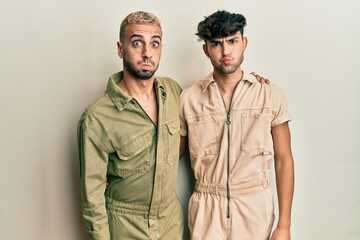 Homosexual gay couple standing together wearing casual jumpsuit puffing cheeks with funny face. mouth inflated with air, crazy expression.