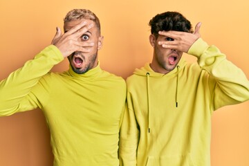 Homosexual gay couple standing together wearing yellow clothes peeking in shock covering face and eyes with hand, looking through fingers with embarrassed expression.