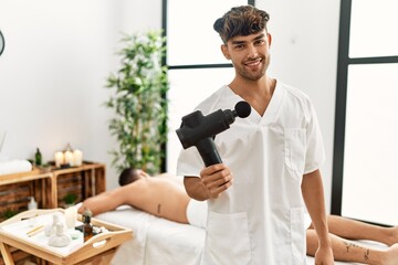 Fototapeta na wymiar Young hispanic man holding therapy massage gun at wellness center looking positive and happy standing and smiling with a confident smile showing teeth