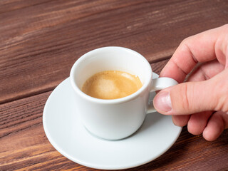close-up of a man's hand holding a small white cup of aromatic espresso over a white saucer. Wooden background, side view.